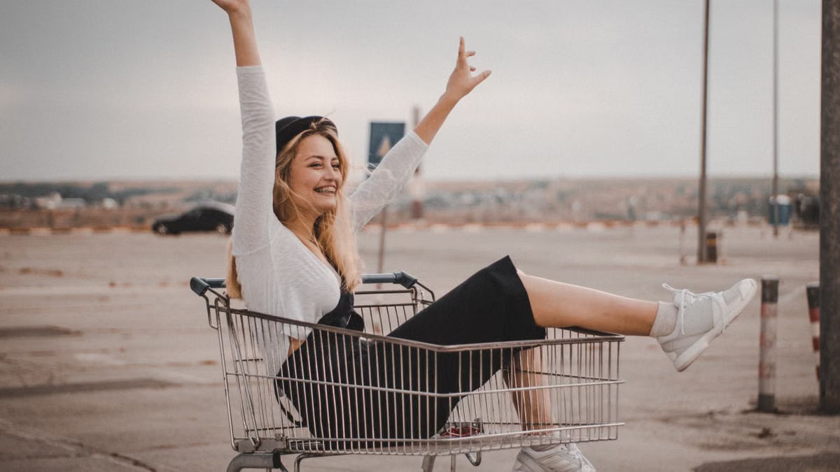 Happy woman rides shopping cart with hands raised, enjoying a playful moment outdoors.